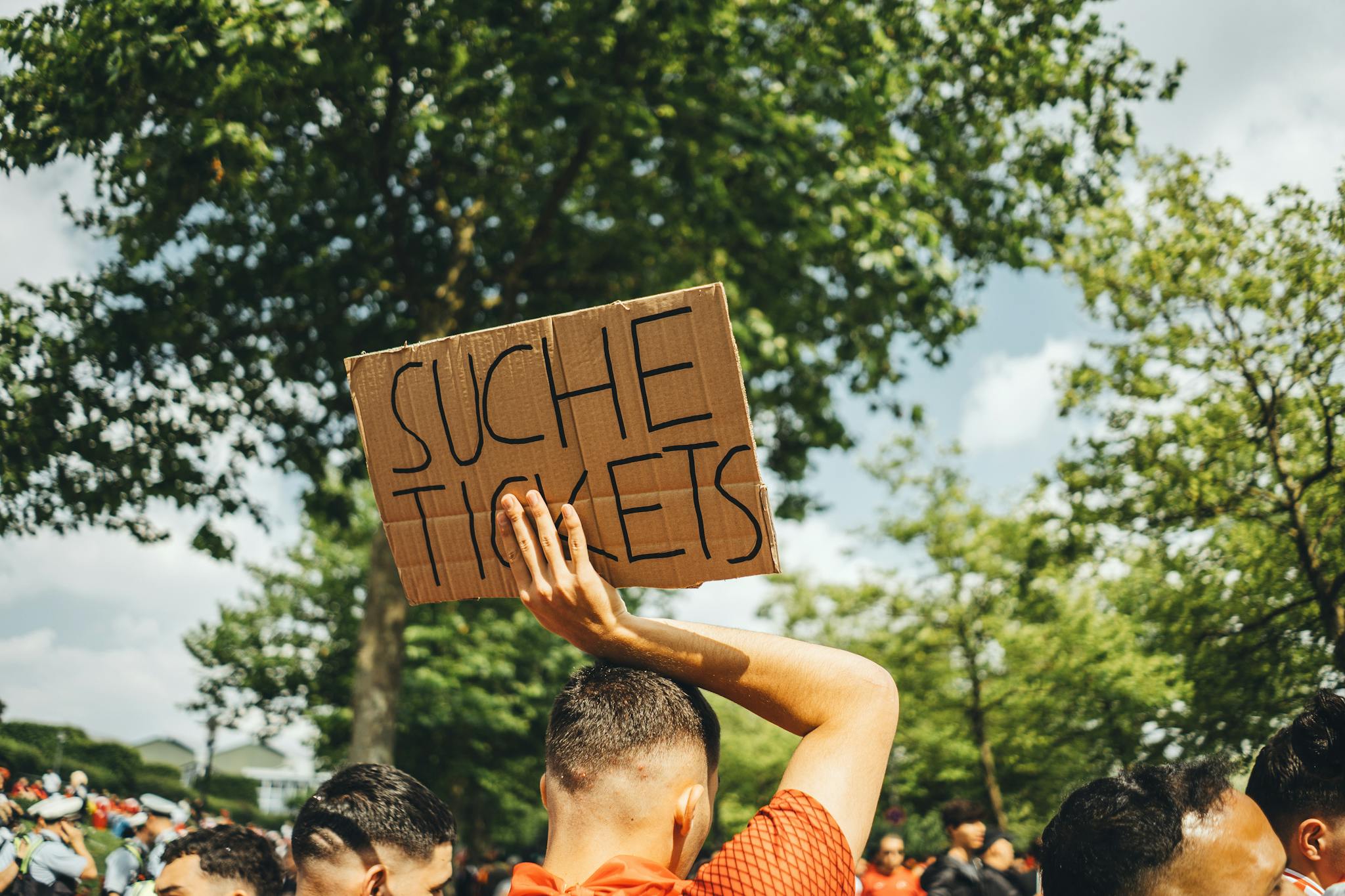 A man in a crowd holds a sign reading 'Suche Tickets' at an outdoor event, seeking extra admissions.